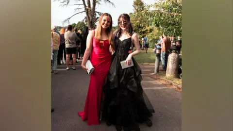 Paul Brewer Ellie in a black dress smiling and looking at the camera stood next to her friend in a red dress. Both girls look very happy and are stood outside on their prom day.