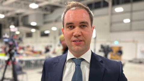 A man with short brown hair, a blue suit and tie looking at the camera