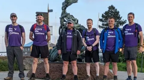 Six men stand in a row and are all wearing purple t-shirts which say 'Every Life Matters'