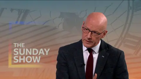 John Swinney, wearing glasses and a suit with burgundy tie, sits in front of a The Sunday Show background.