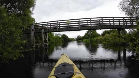 A kayaker's view showing the front of a yellow kayak approaching a wooden footbridge in the middle of a dark, wide river lined by trees