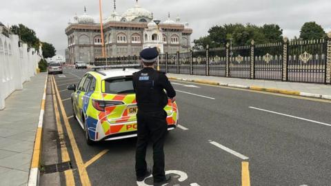 A policeman stands by a car at the gated entrance to Gravesend Gurdwara
