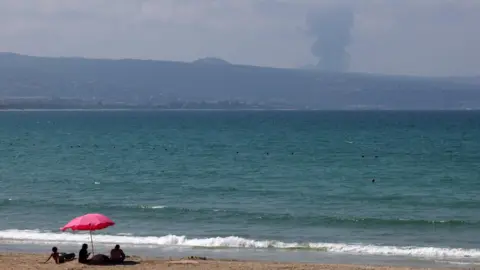 A family sits on a beach in Tyre, Lebanon, as smoke rises on the Lebanese side of the border with Israel during heavy exchanges of fire between Hezbollah and the Israeli military on 25 August 2024