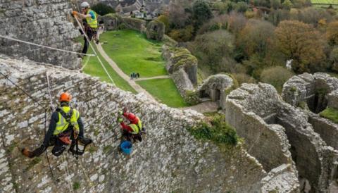 A view from above of three people on abseiling ropes hanging from the castle ruins. They are wearing hard hats and hi-vis vests. In the background is a village and some woods