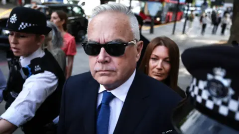 Former BBC news presenter Huw Edwards, wearing a navy suit, blue tie and sunglasses, is flanked by police officers outside a London court in July