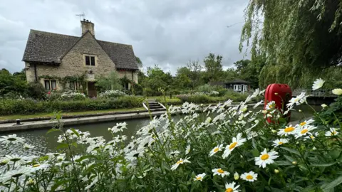 EstherJ FRIDAY - A grey stone cottage sits on the banks of the canal at Iffley Lock. In the foreground there are large white and yellow flowers with an orange life-saving ring. The grey water of the canal runs between them with a willow tree overhanging. Above the sky is grey.