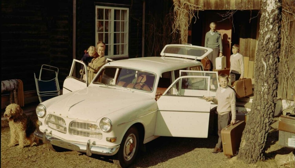 Familie pakker en Volvo stasjonsvogn i 1964. På bildet: en hund, mor, far, tre barn og en hvit bil. Et hus i bakgrunnen.
