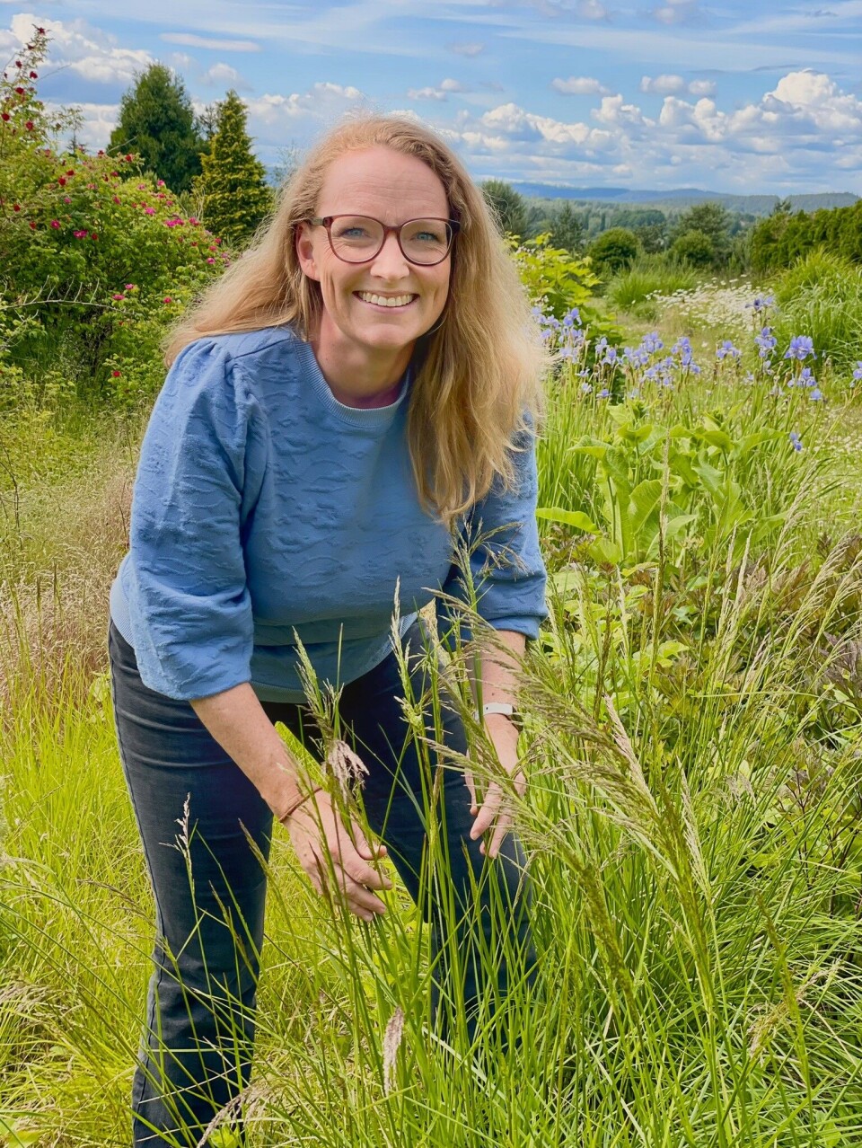 Biolog Camilla Lorange Lindberg står mellom høyt gress. Hun har langt, lyst hår, briller og blå genser. I bakgrunnen ser man blå blomster og høye trær.