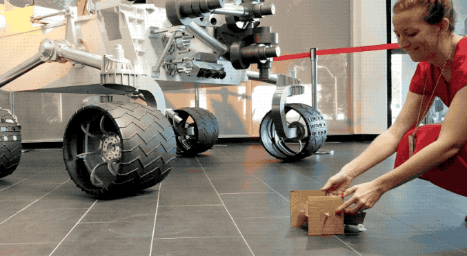 Animated image of a woman releasing a cardboard rover that jumps forward. A model of the Curiosity rover can be seen in the background.