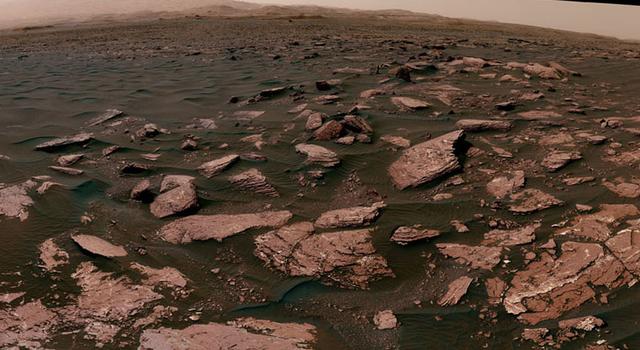 Red rocks and sand cover this Mars landscape with dunes rising in the distance