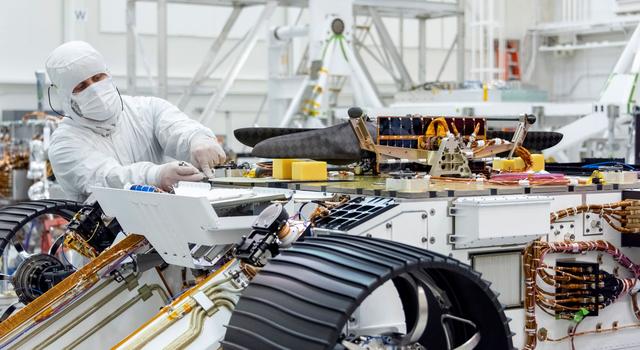 An engineer dressed in white protective gear places the Mars helicopter onto the belly of the rover.