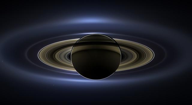 An image of Saturn lit up from behind by the Sun with the planet's moons, Earth and Mars visible as tiny specs of light in the background..