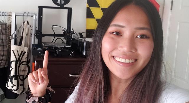 Christine wears a scrunchy on her wrist while pointing to the 3D printer, which sits on a dresser between a rack of clothes and a flag hanging on the wall.