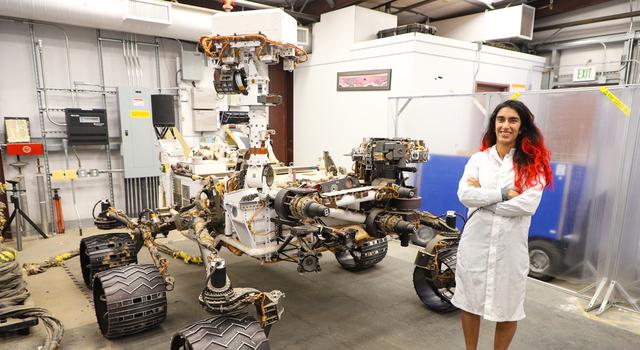 Farah Alibay, wearing a white lab coat, poses for a photo in front of an engineering model of the Curiosity rover