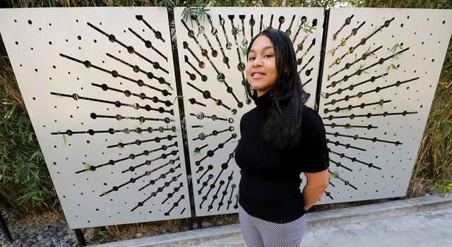 Mariah Woody poses for the camera with her hands clasped behind her back in front of a metal starburst screen.