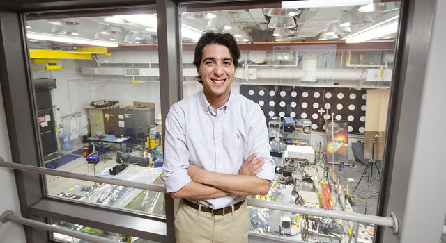 Max Rudolph crosses his arms and smiles at the camera standing in front of a glass window that looks down on the In-Situ Instrument Laboratory