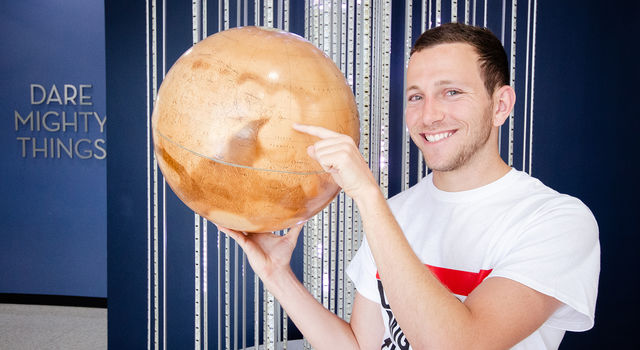 Miles Fertel smiles at the camera while holding a Mars globe in one hand and pointing to Mars 2020's planned landing spot with the other hand. He's standing in front of a light sculpture and a sign that says "Dare Mighty Things."