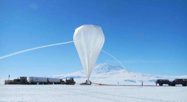 A large tear-drop shaped balloon towers above surrounding work trucks on a flat expanse of snow.
