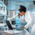 Young Hispanic scientist wearing a lab coat, looking under microscope while using laptop in a laboratory.