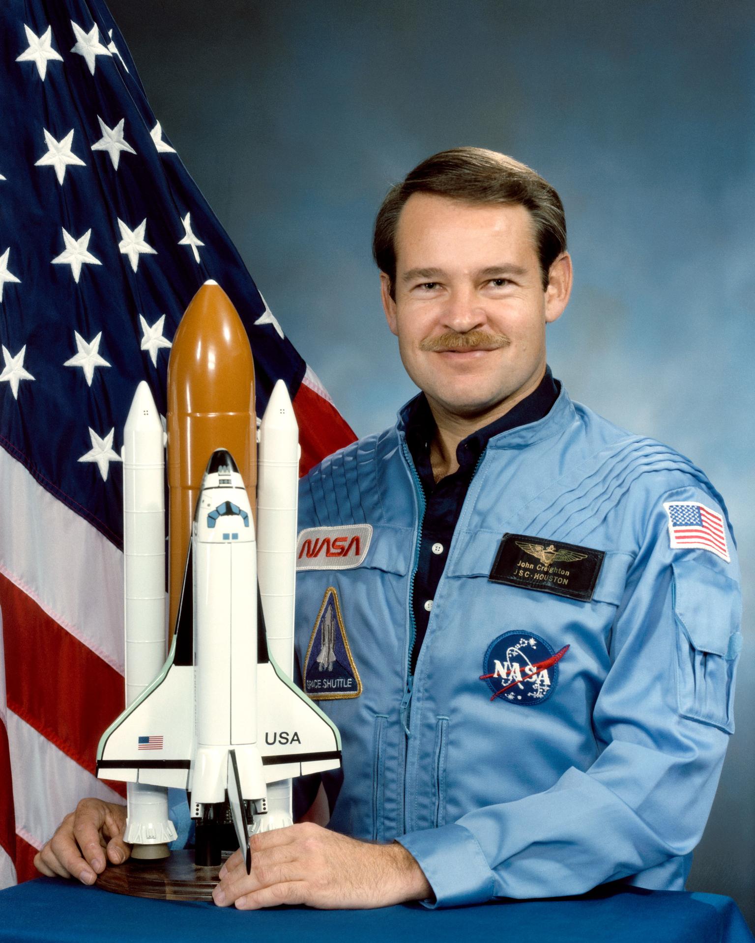 A man smiles, holding a rocket model before him. He is wearing an astronaut jumpsuit and stands before the American flag.