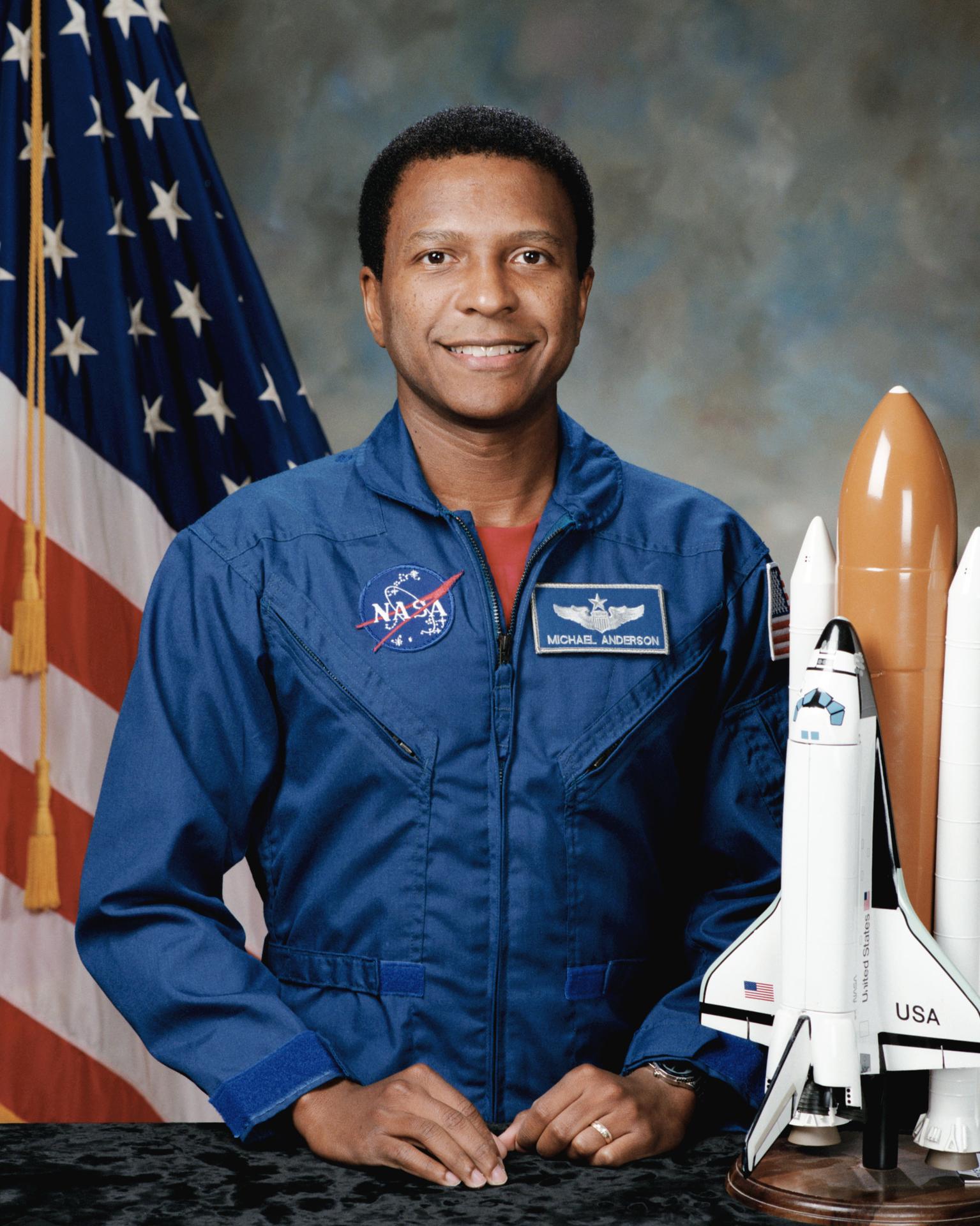 A man stands before the American flag, with a rocket ship model at the side