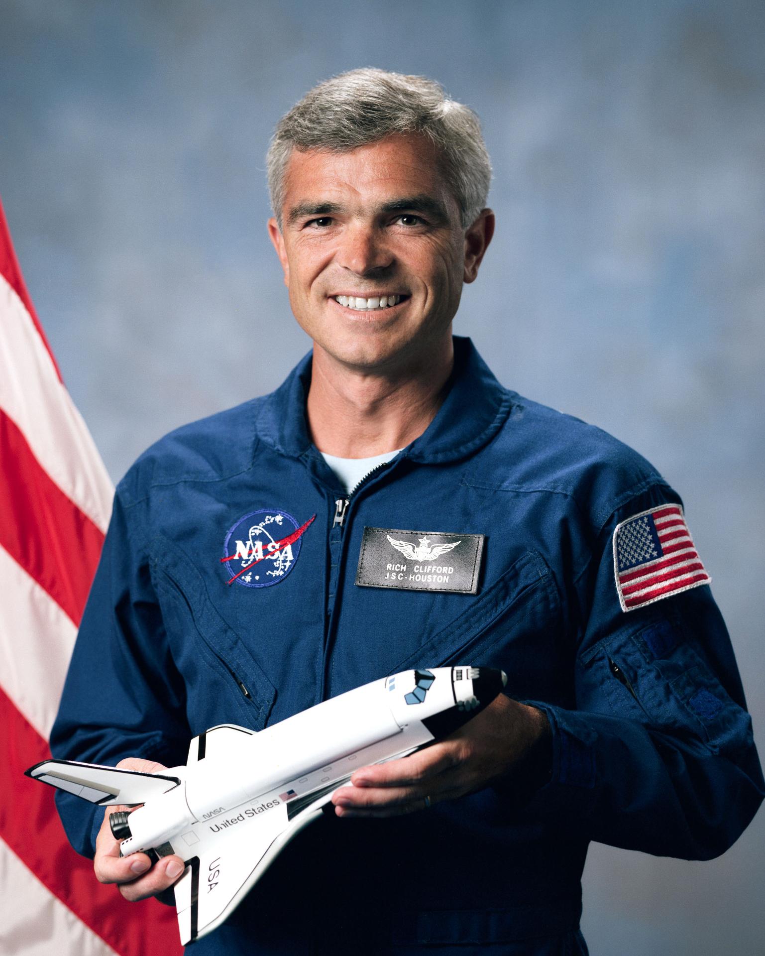 A man stands in front of the American flag, smiling and holindg a model rocket in his hands before himself.