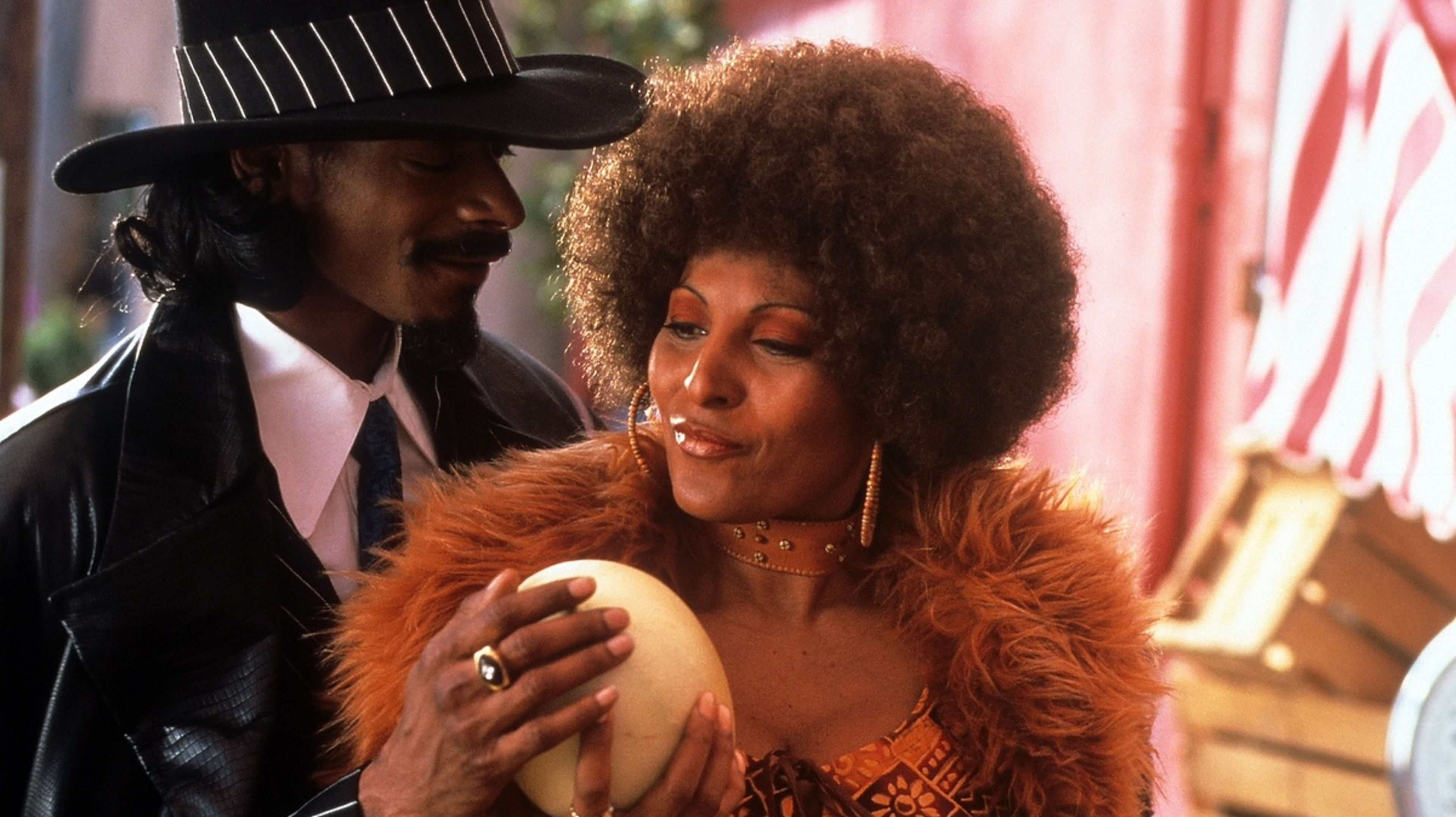 Snoop Dogg and Pam Grier share a moment; Snoop Dogg in a pinstripe hat and leather coat, Pam Grier in a fur-trimmed coat, afro hairstyle, holding a large egg