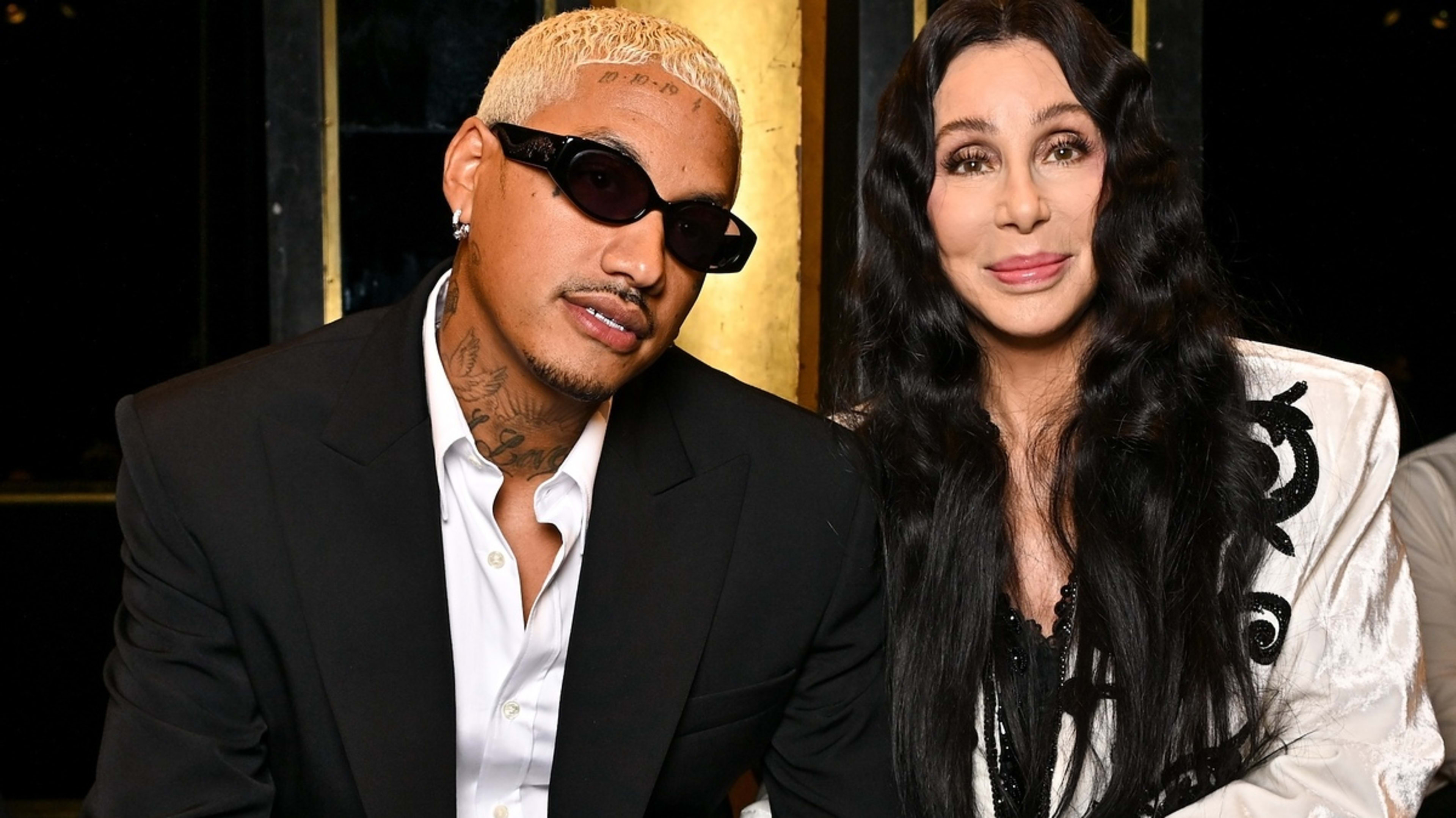 Cher and Alexander Edwards are seated together, with Cher wearing a stylish white jacket adorned with black details and Edwards in a black suit and sunglasses