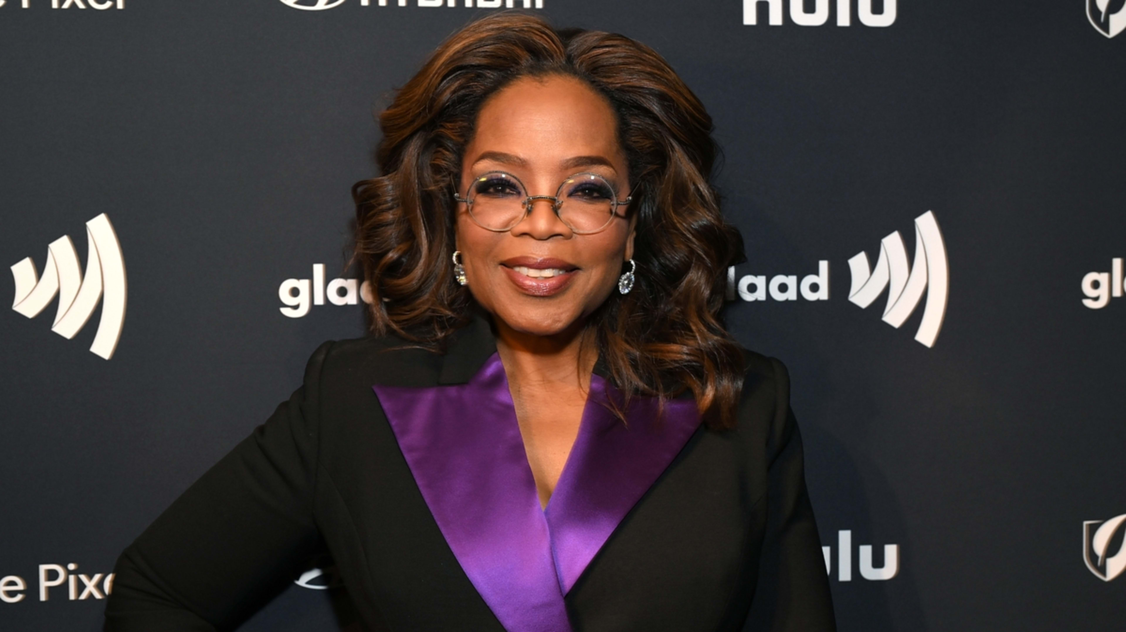 Oprah Winfrey at a GLAAD event in a stylish black outfit with a purple lapel. She is wearing glasses and smiling at the camera