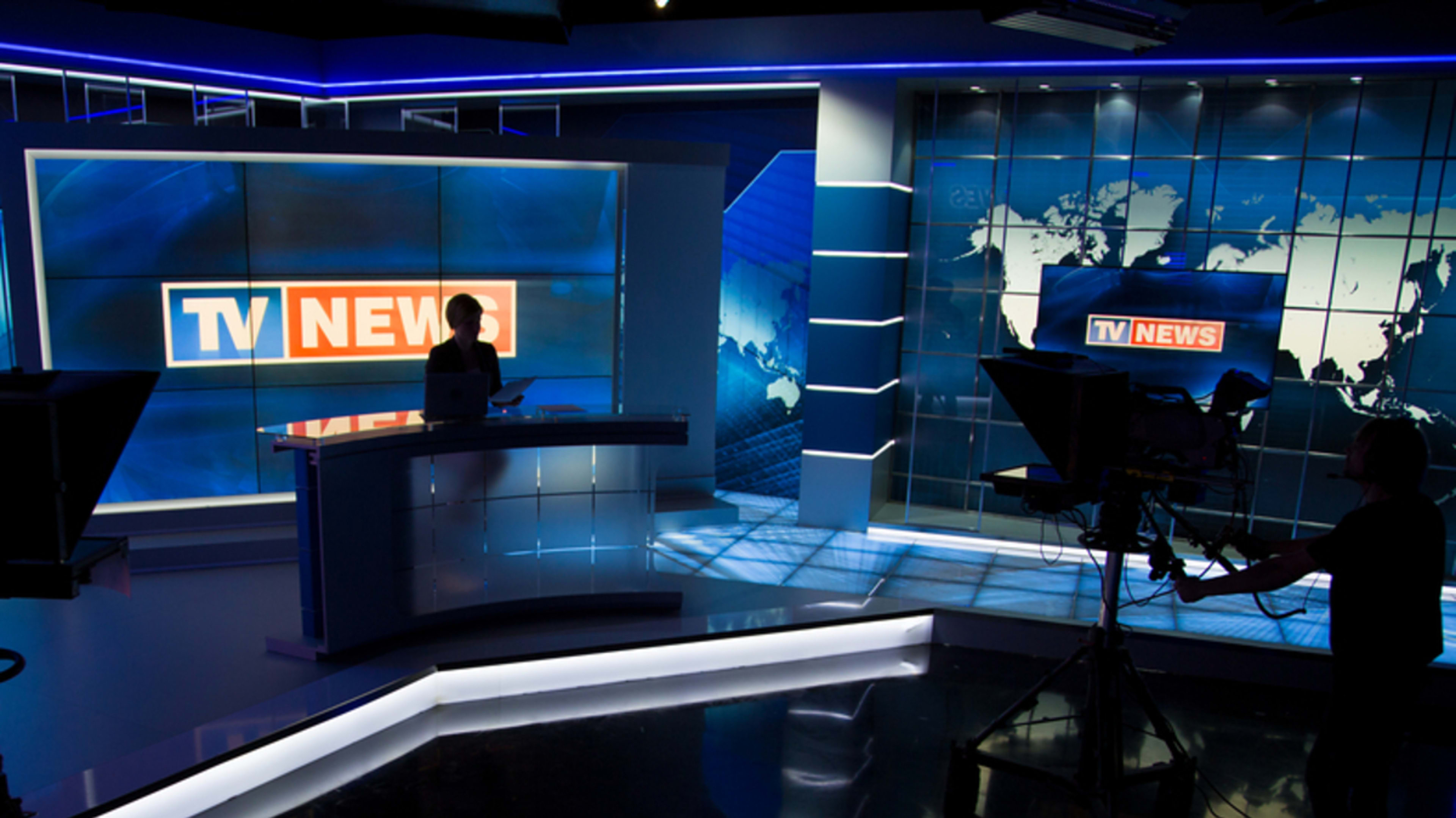 A TV news studio with a news anchor at the desk. Camera operators are setting up cameras. The background has a world map and a "TV News" logo