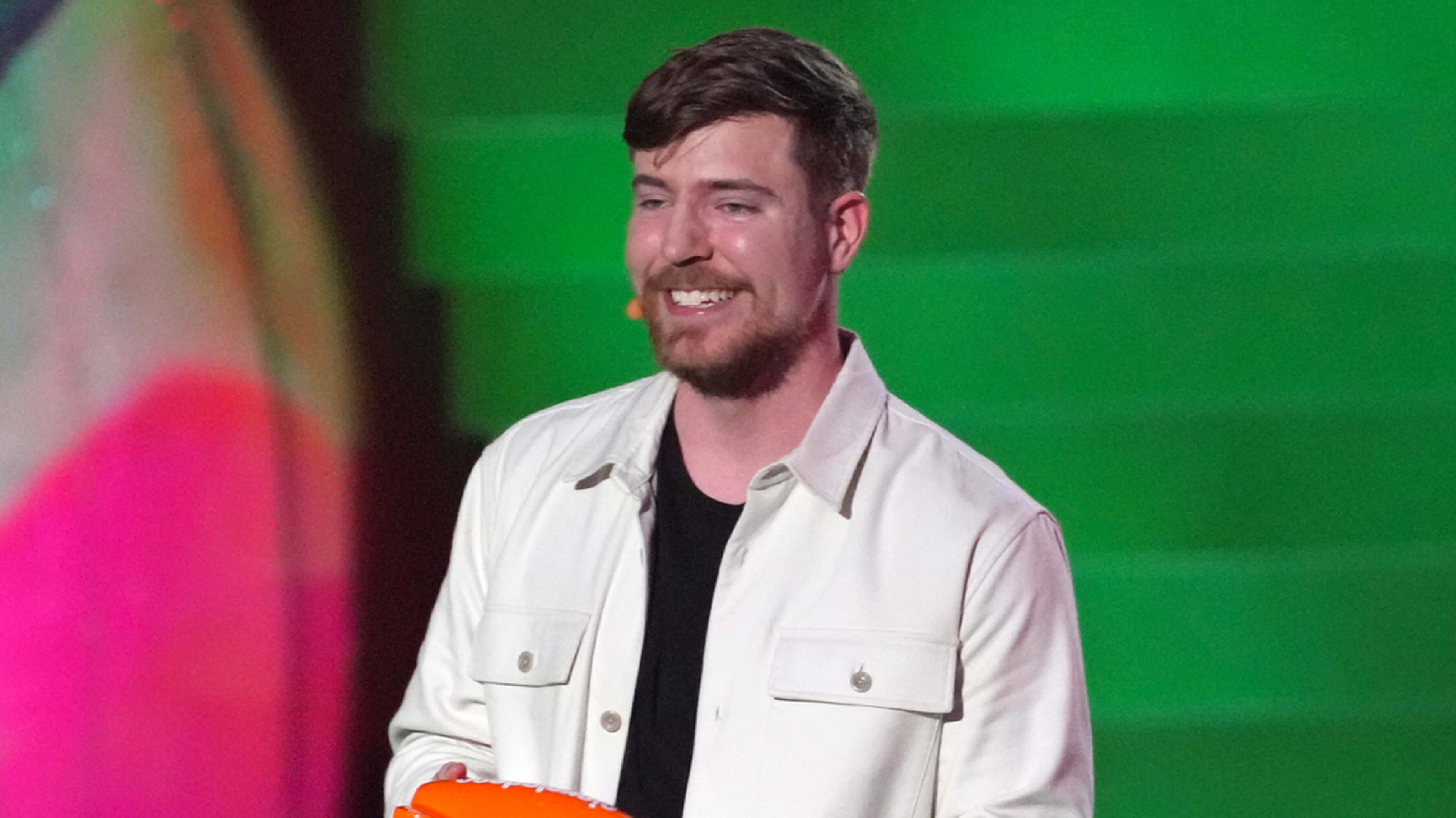 MrBeast, wearing a white jacket over a black shirt, smiles while holding an award at a public event