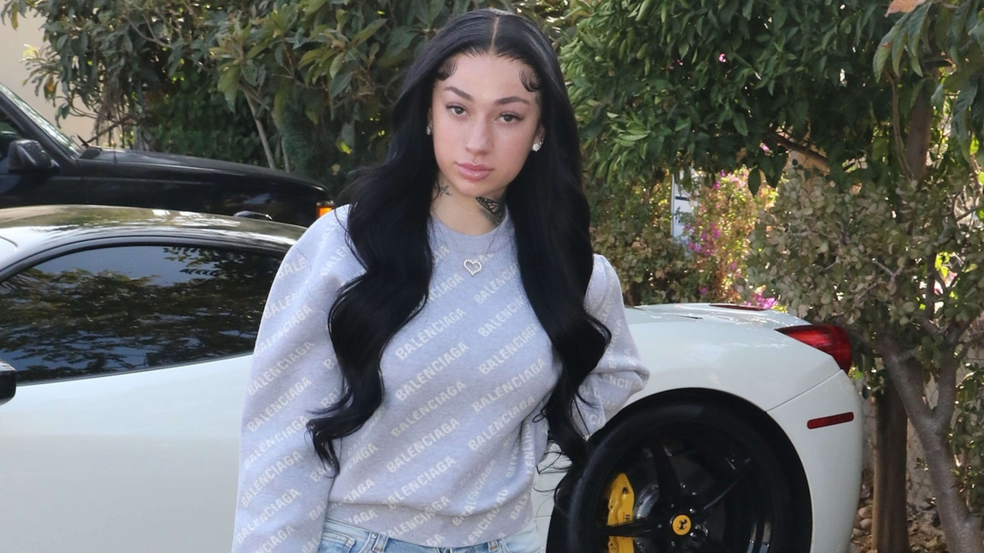 A person with long wavy hair wearing a Balenciaga sweatshirt and jeans stands outside next to a white car. Trees and greenery are in the background