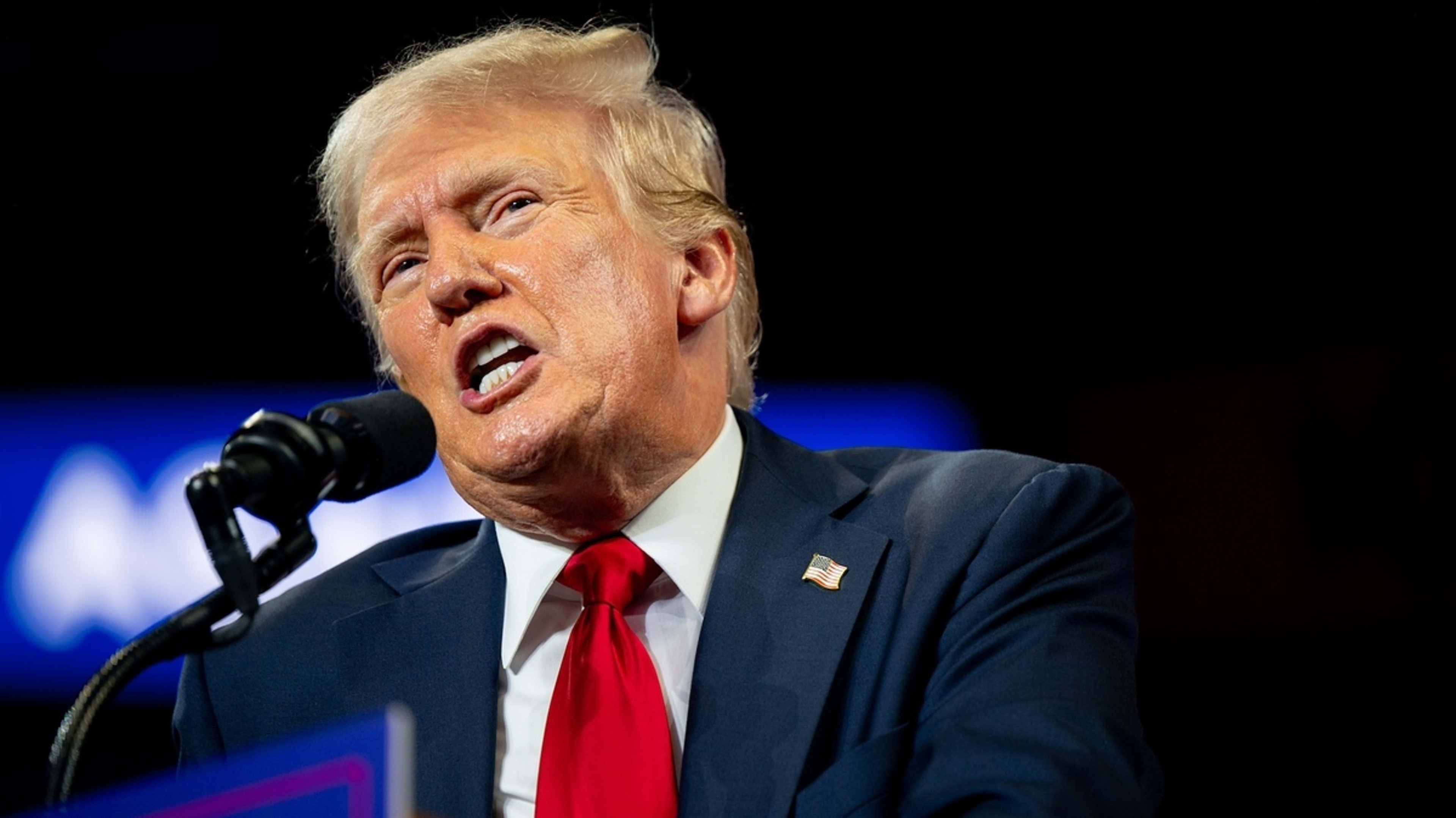 Donald Trump speaks into a microphone at a public event, wearing a suit, white shirt, and red tie