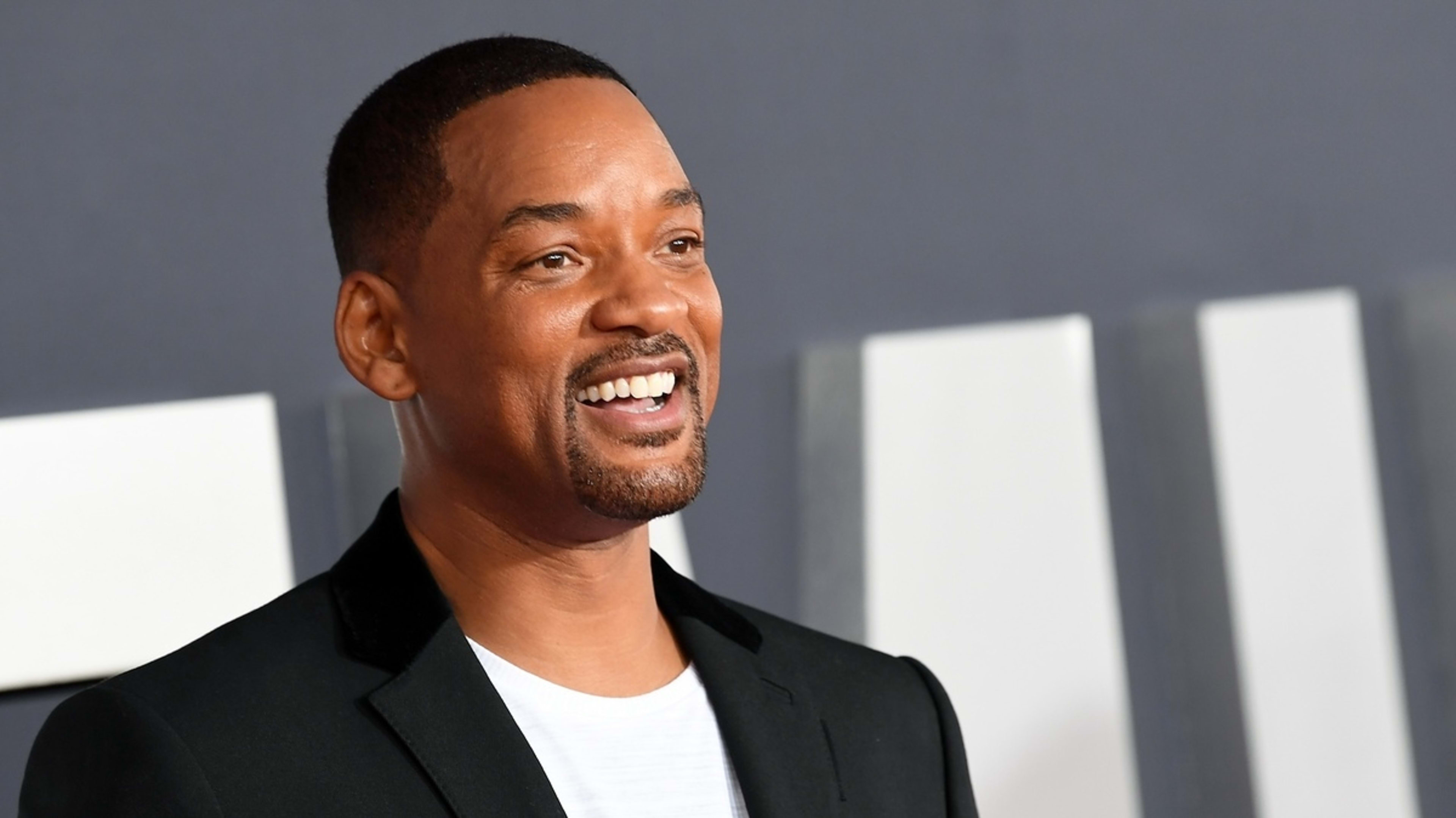 Will Smith dressed in a casual white shirt and black jacket, smiling at an event