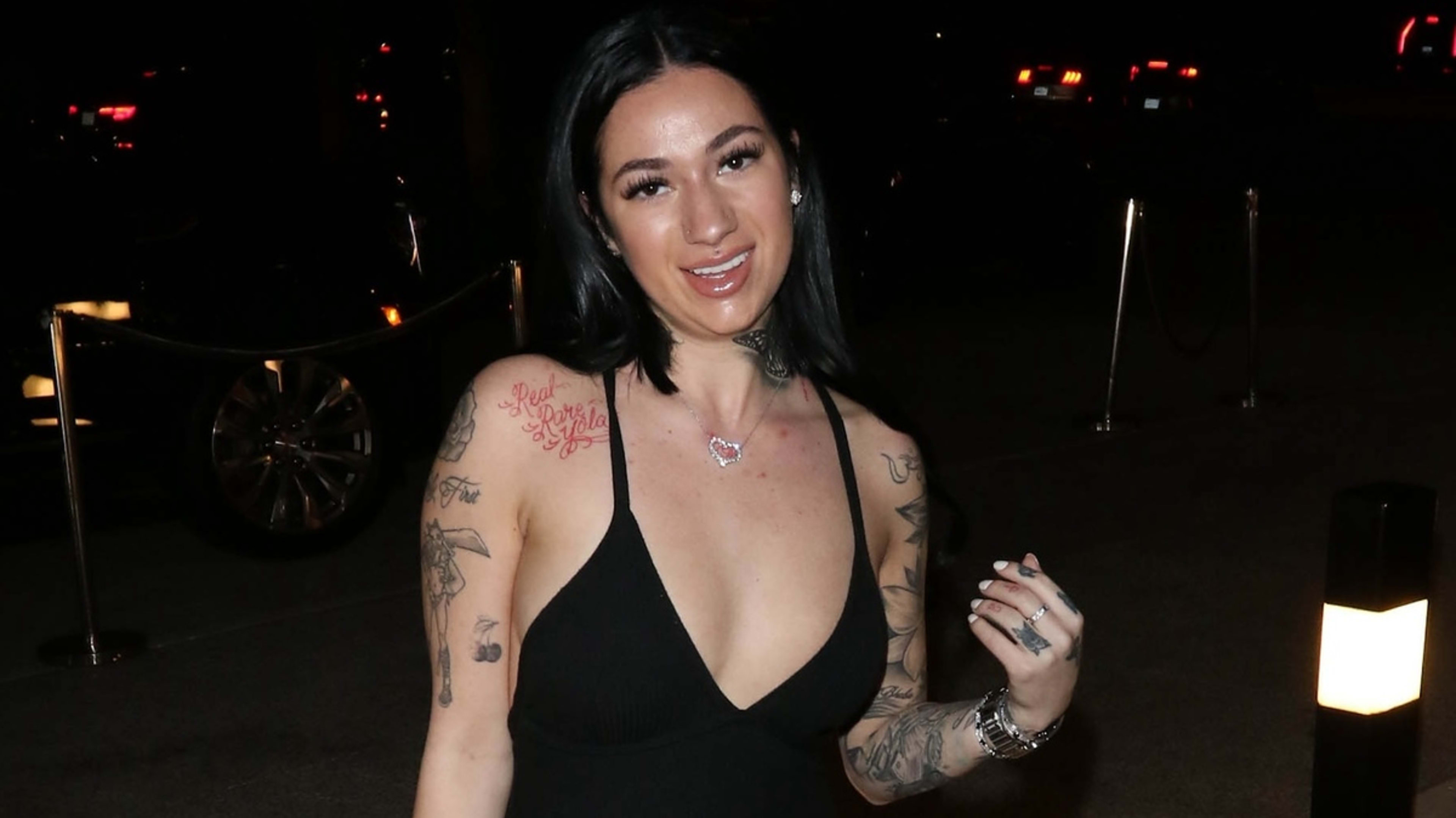 Woman with dark hair and tattooed arms in a sleeveless dress smiles at the camera in an evening outdoor setting