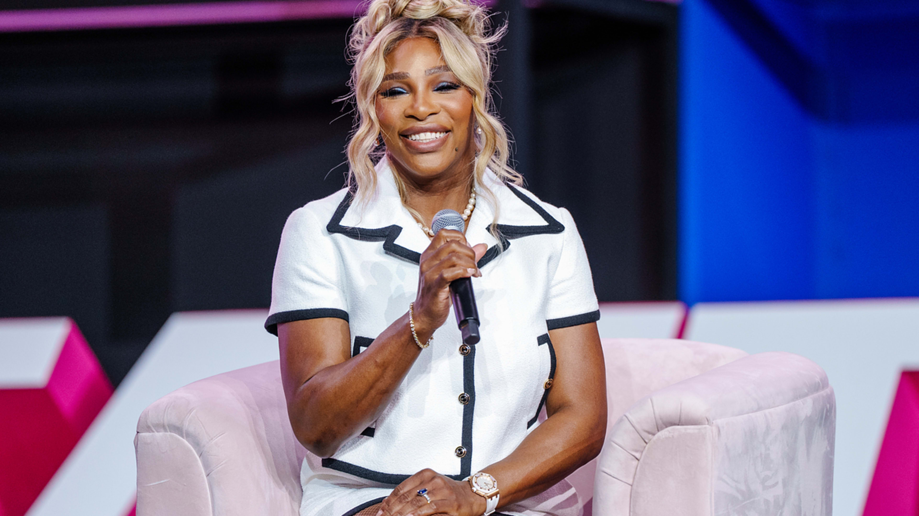 Serena Williams smiles while holding a microphone, dressed in a chic white outfit with black trim, seated on a stage during a sports event interview