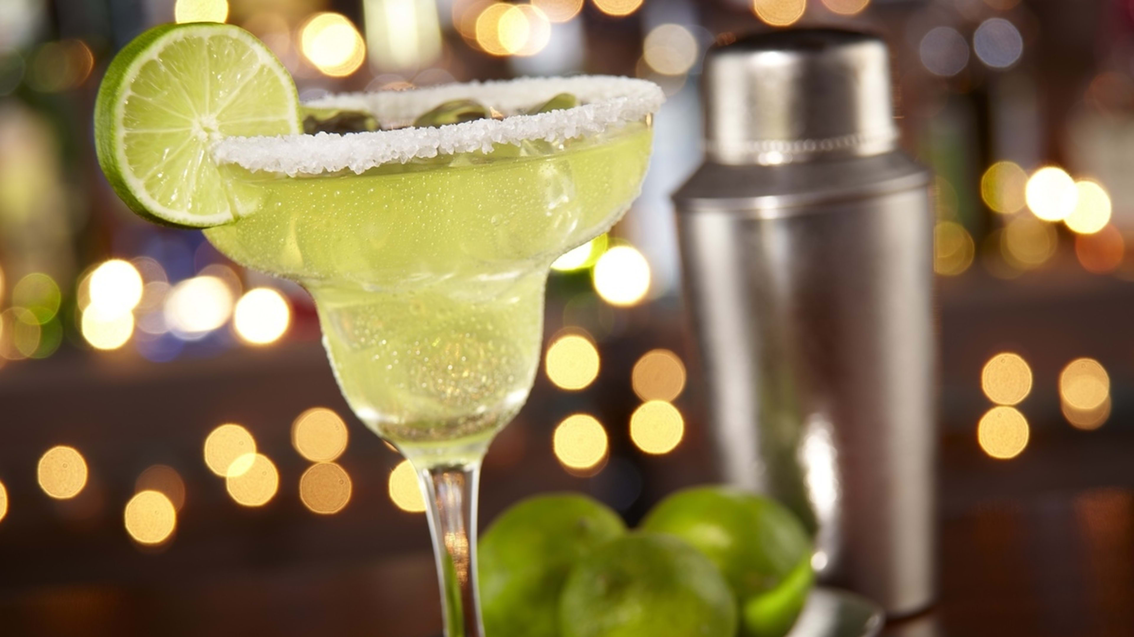 Margarita cocktail with a salt-rimmed glass, garnished with a lime slice. A cocktail shaker and a few limes are in the background