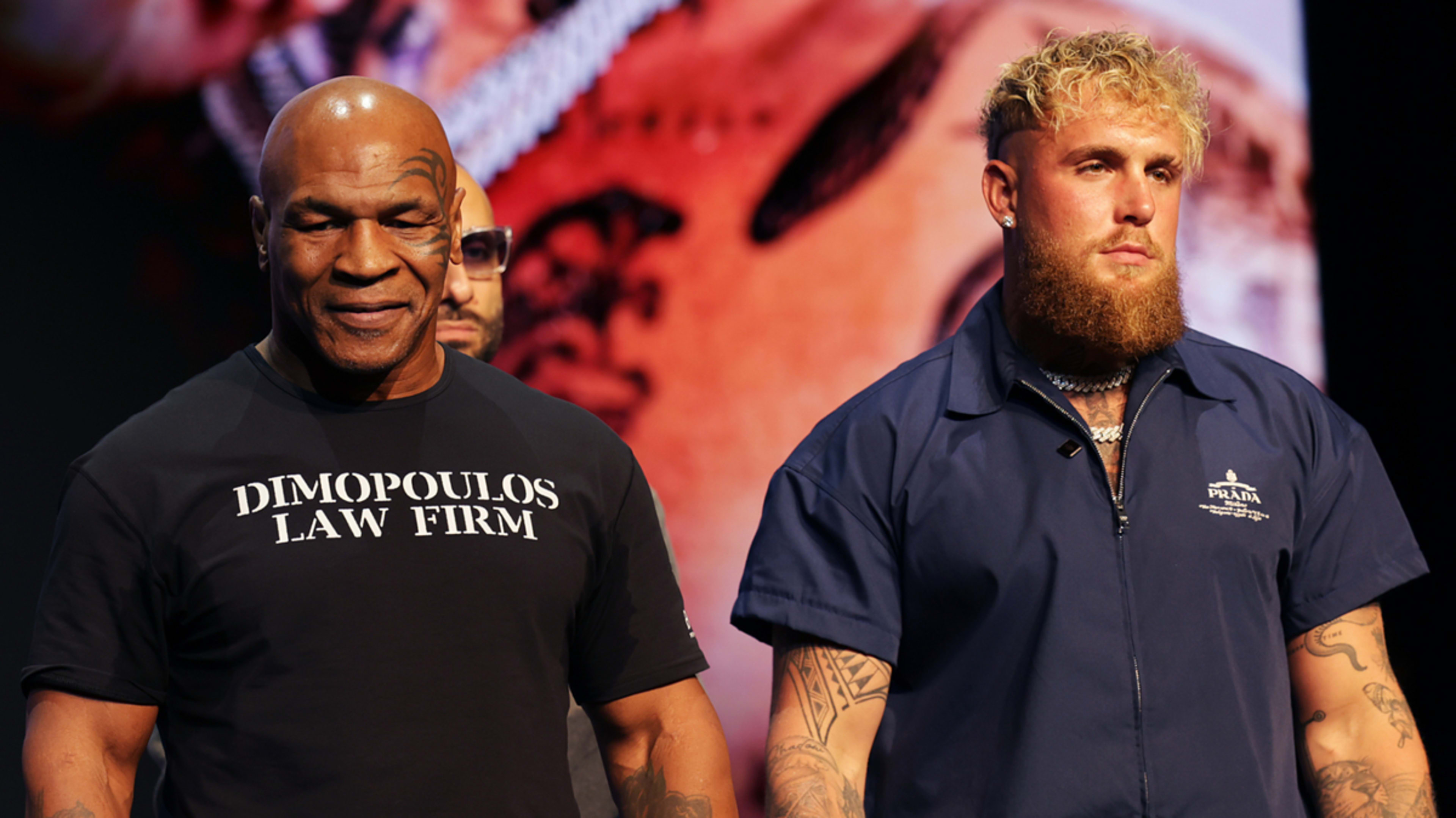 Mike Tyson (left) in a "Dimopoulos Law Firm" t-shirt and Jake Paul (right) in a dark zip-up jacket, posing at an event