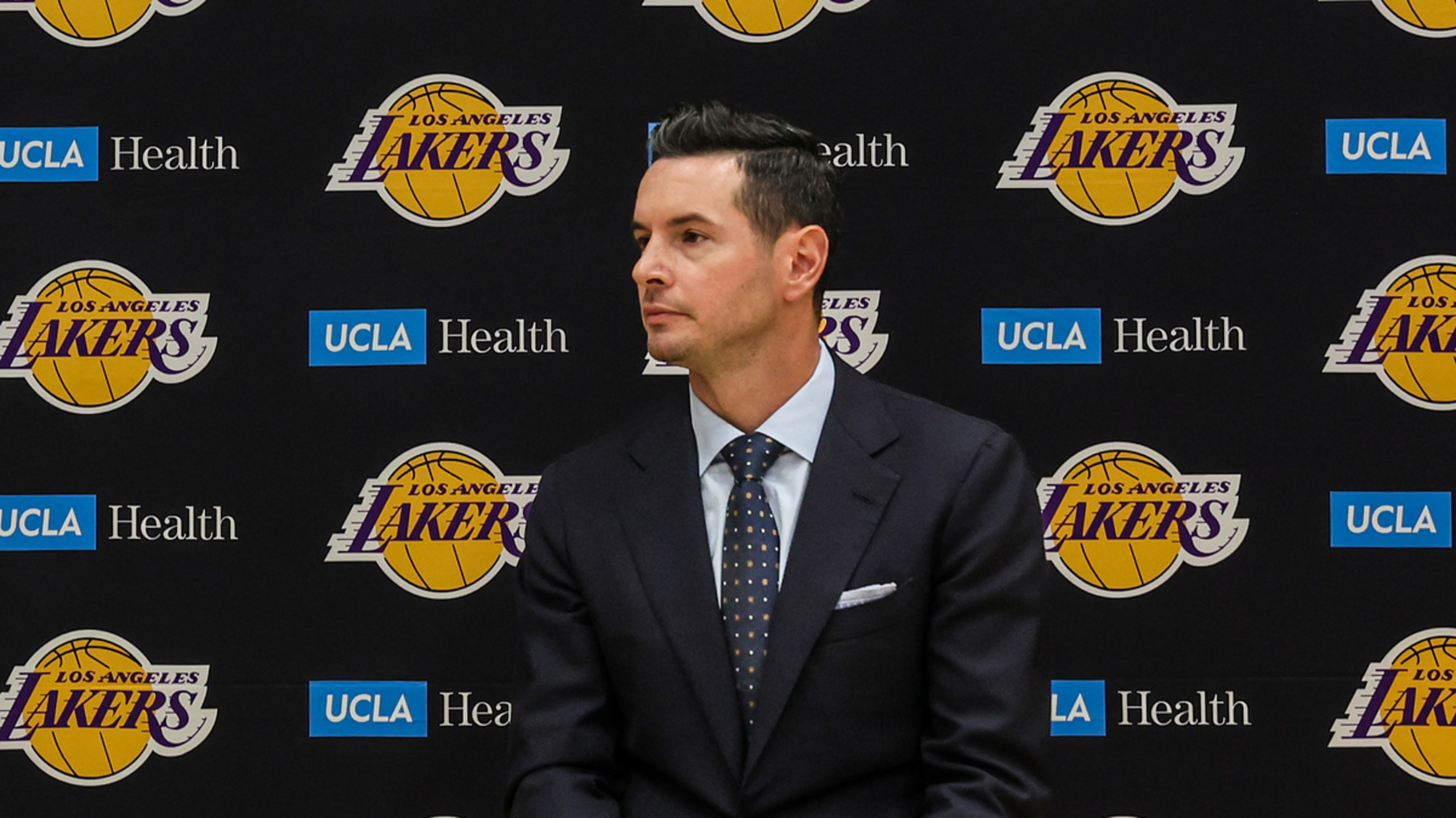 Rob Pelinka speaking into a microphone and Darvin Ham seated at a Los Angeles Lakers press conference backdrop with sponsor logos