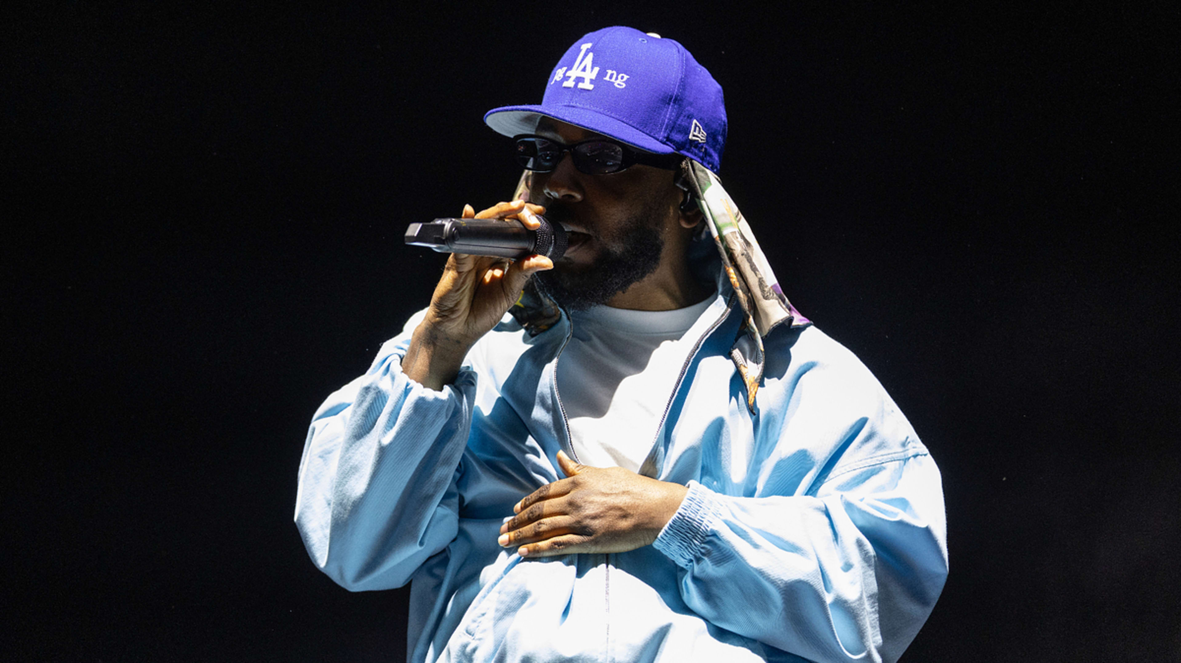 A person wearing a blue jacket, white shirt, blue LA cap, and sunglasses sings into a microphone on stage