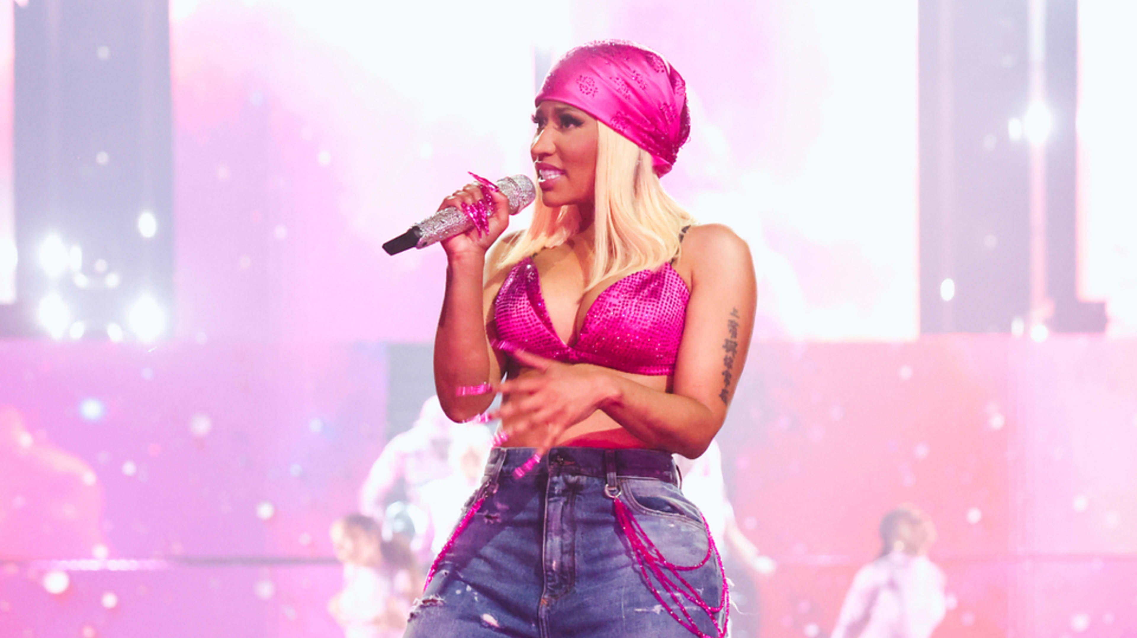 Cardi B performs on stage in a pink top, ripped jeans, and a pink headscarf, holding a microphone. Pink, ethereal background and backup dancers visible