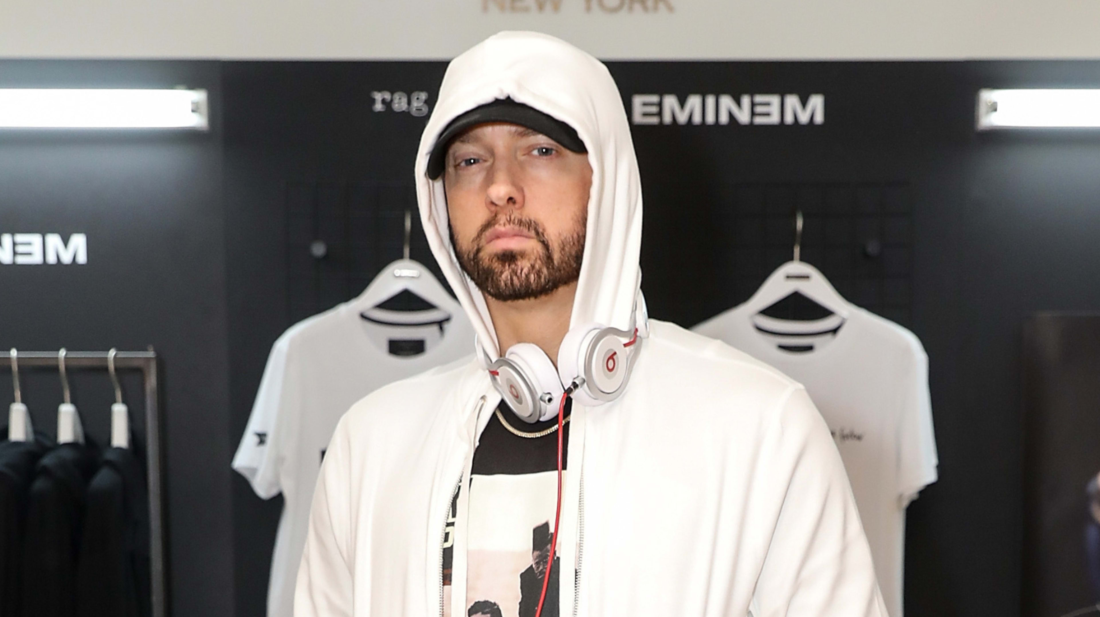 Eminem wearing a hooded jacket and headphones stands in a store with his merchandise