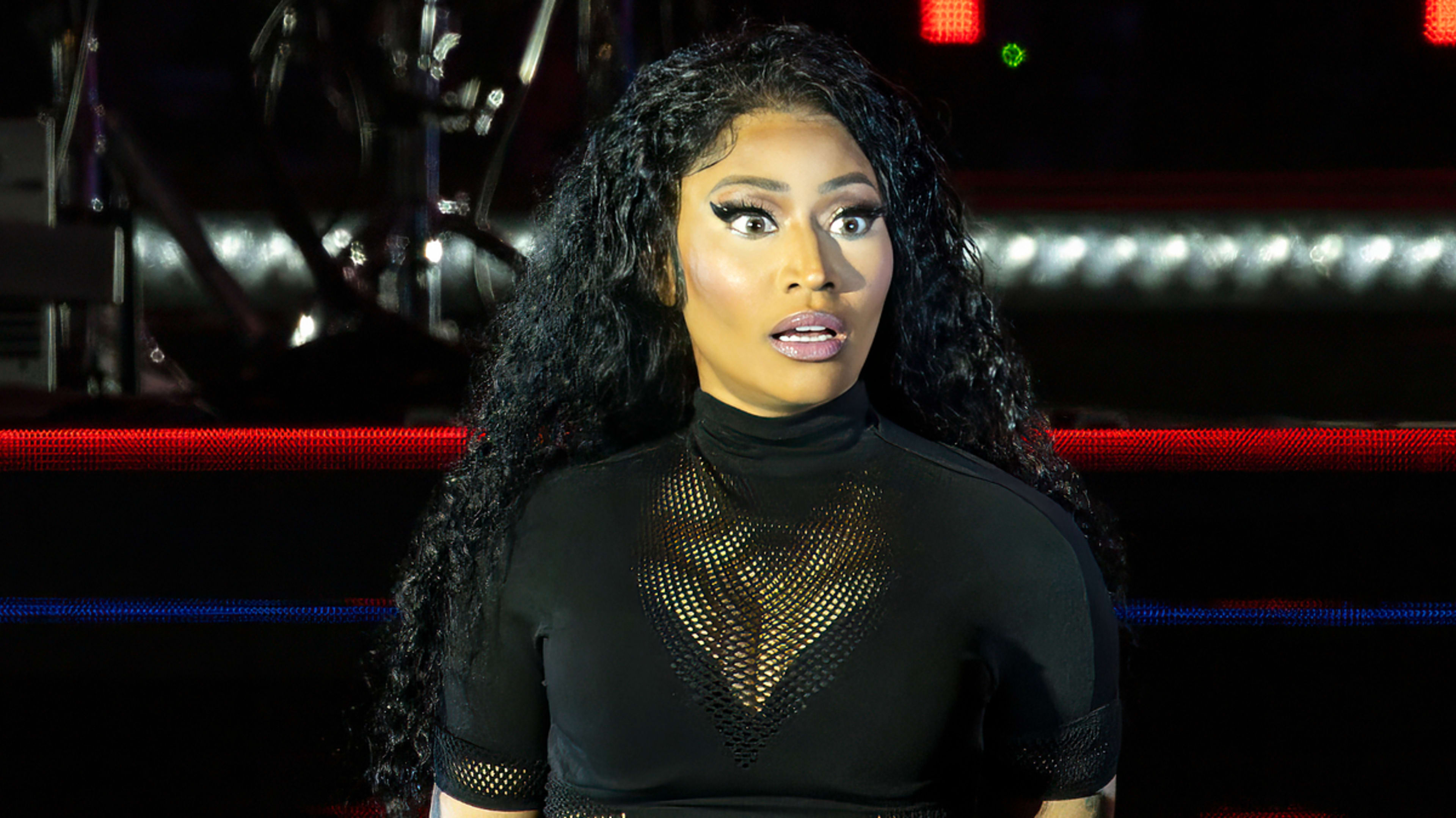 Nicki Minaj, in a black mesh outfit, performs on stage, looking surprised with wide eyes and open mouth. Studio lights and equipment are visible in the background