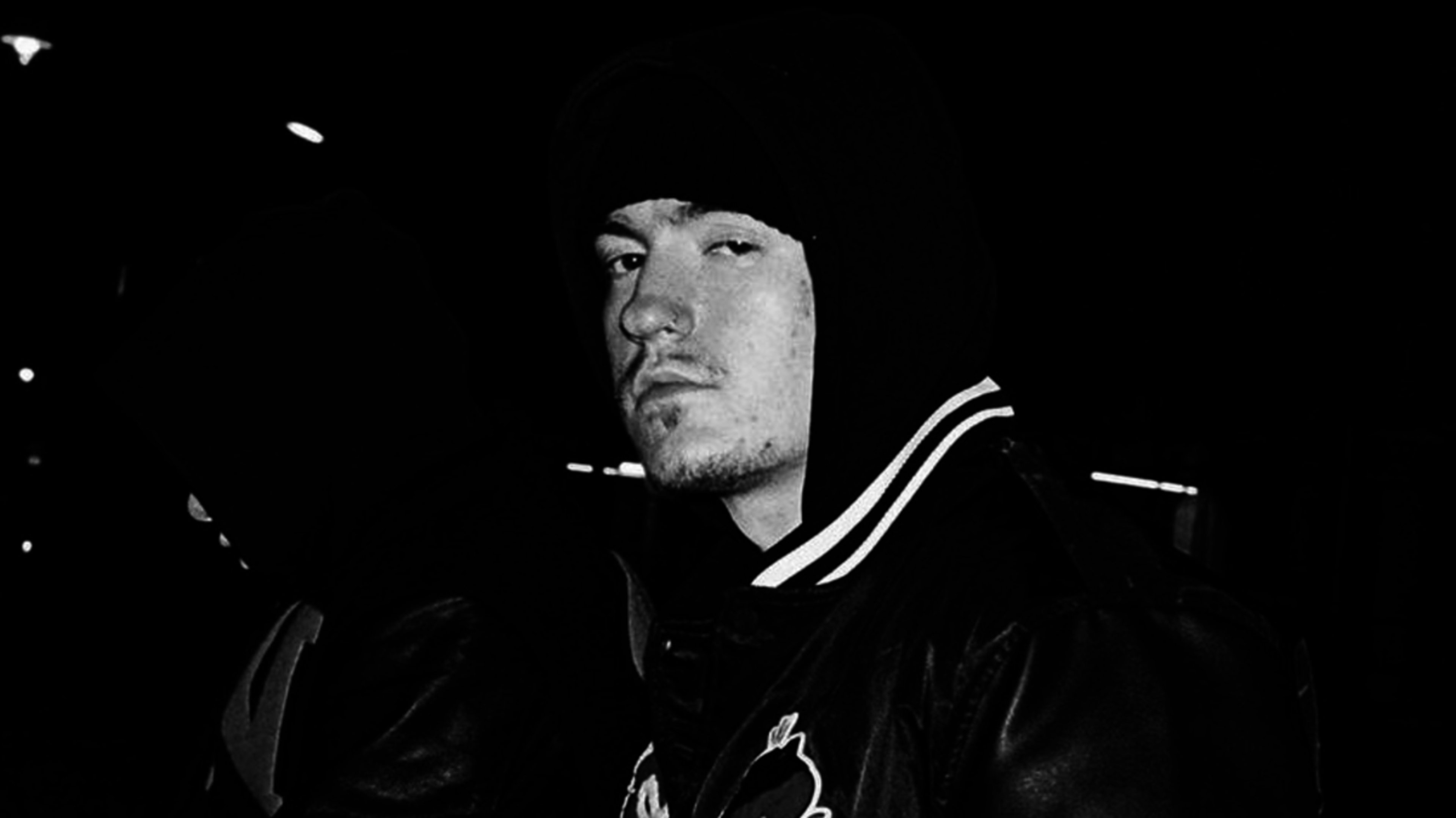 A man wearing a hooded jacket and a beanie at night. He has a serious expression and is looking towards the camera. His identity is not known