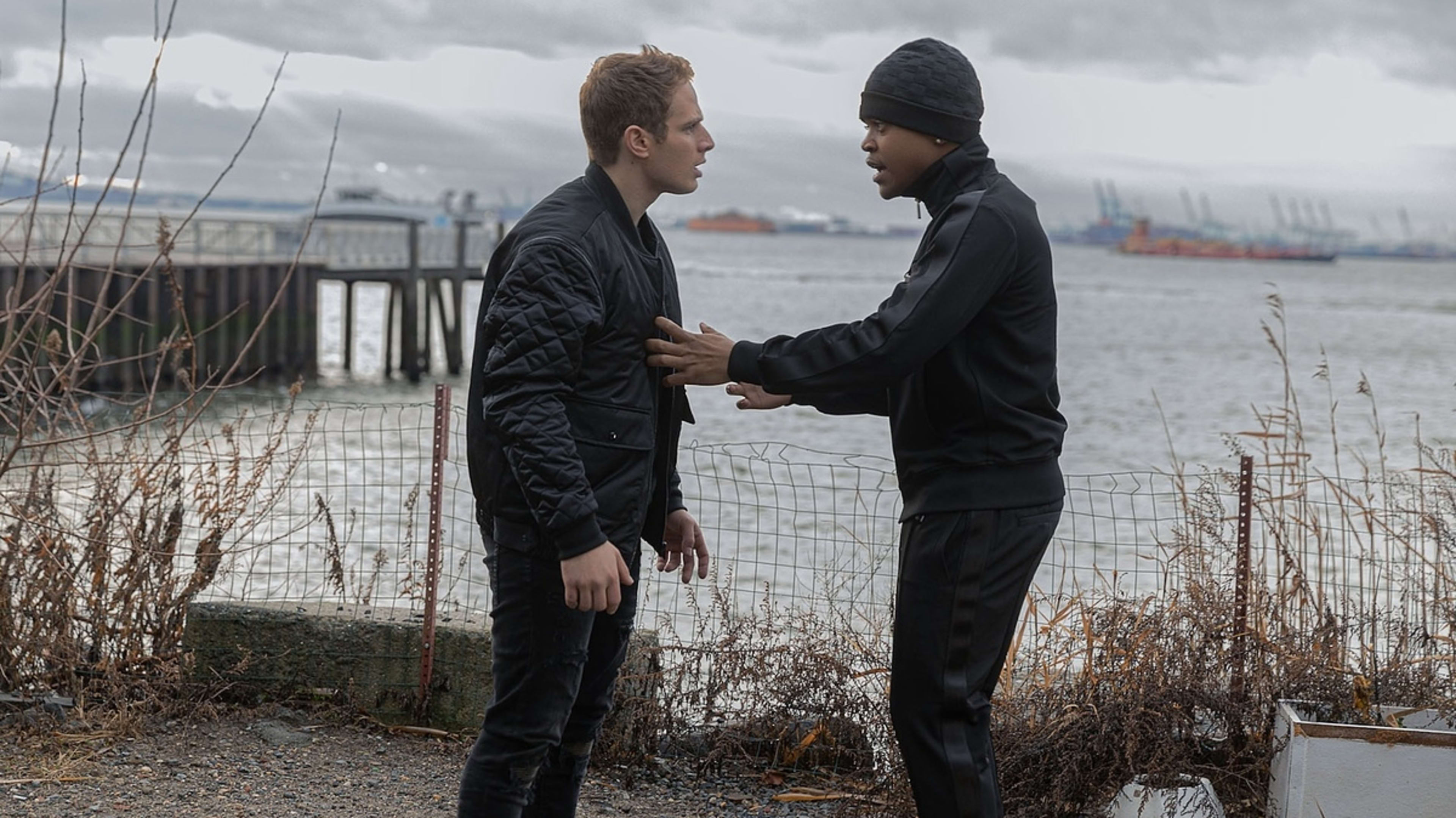 Two characters from a film engage in a tense conversation by a waterfront with industrial ships in the distance