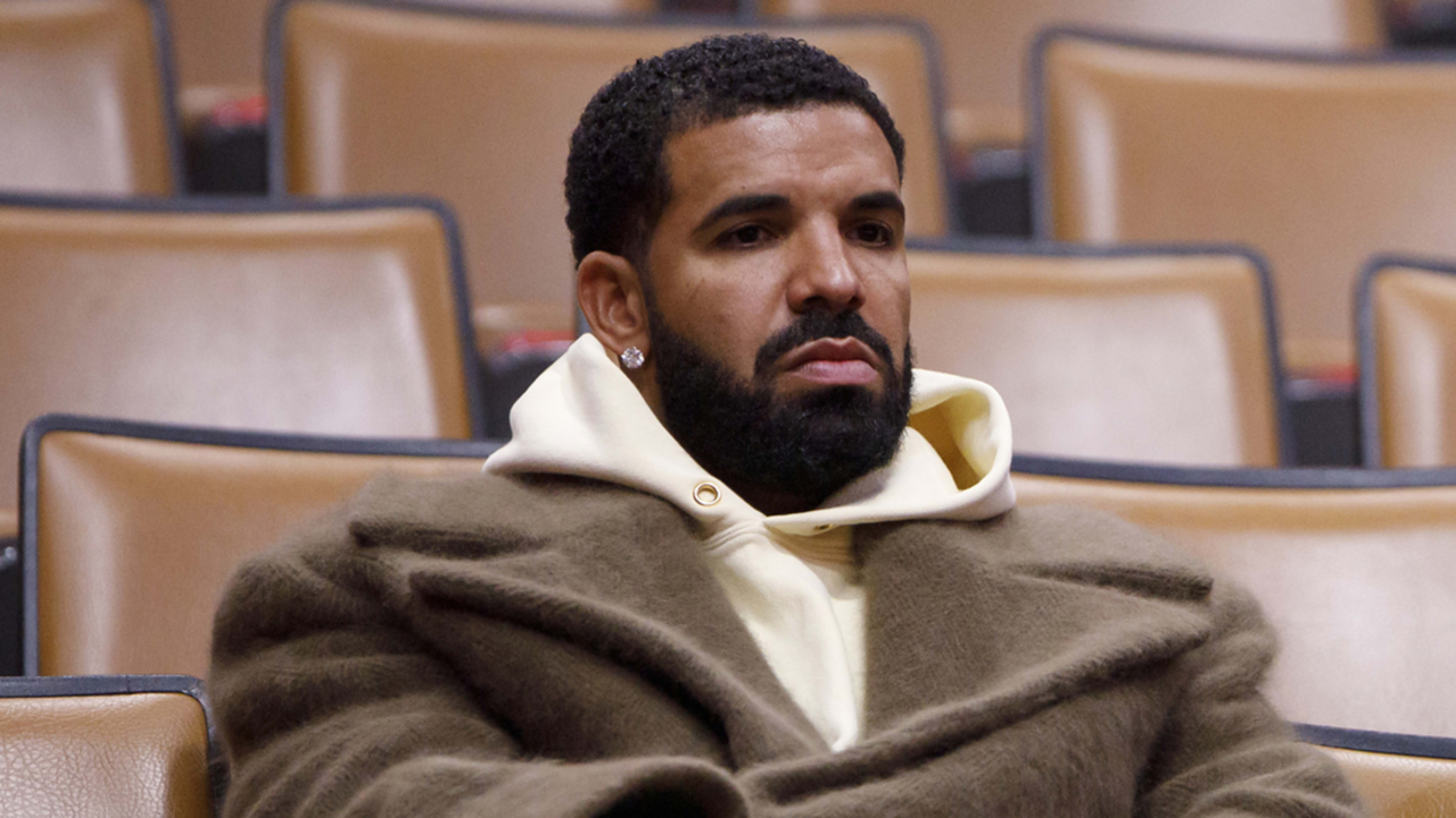 Drake sits alone in an auditorium wearing a large coat with a thoughtful expression