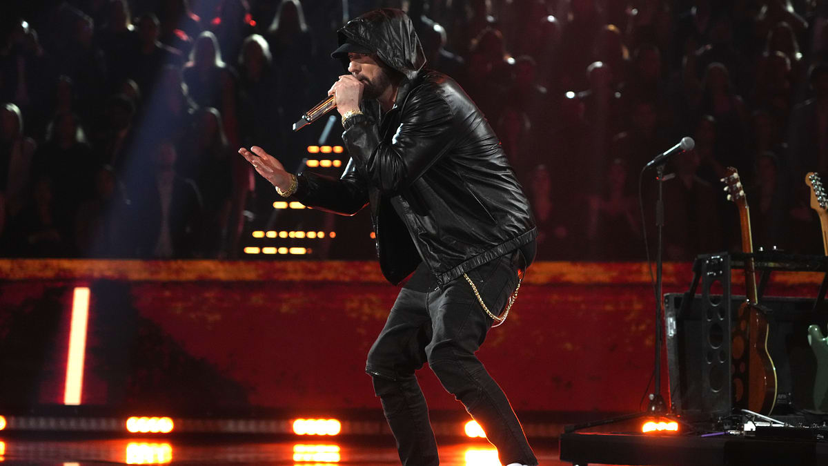 Musician in leather jacket and hat performing onstage with microphone, background has red lighting and audience silhouette