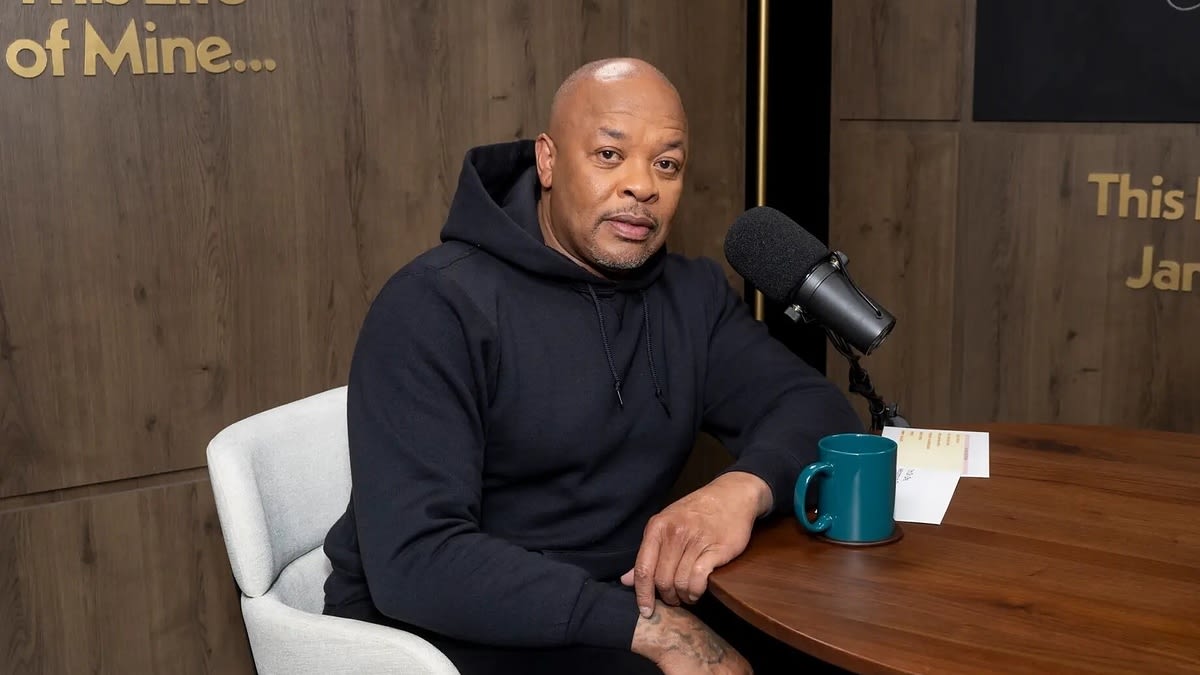 Dr. Dre sits at a podcast desk with a microphone and a mug. He wears a black hoodie