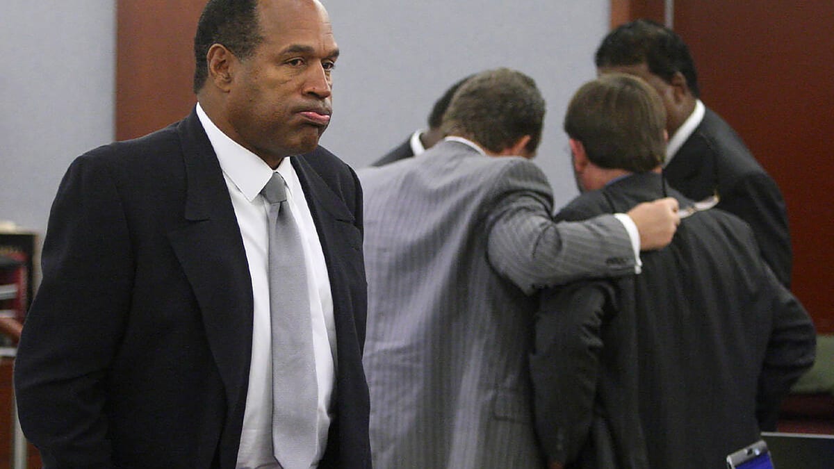 O.J. Simpson stands in a courtroom wearing a dark suit and white shirt, while three men in suits huddle in the background