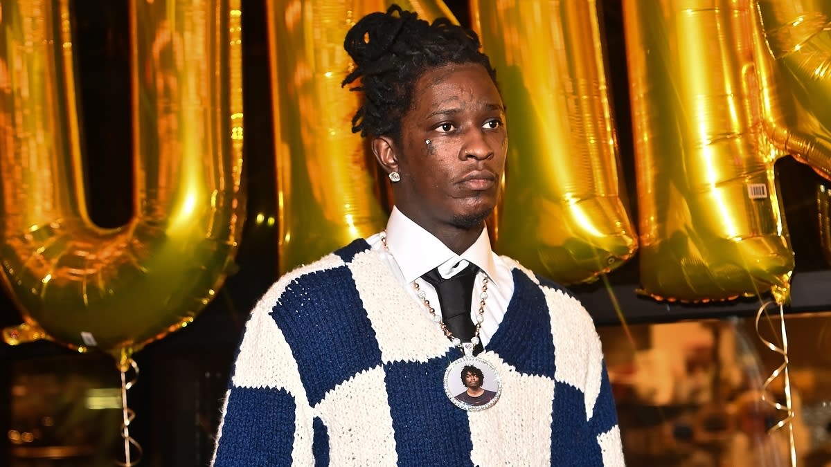 Young Thug wearing a blue and white checkered sweater, a white shirt, and a black tie, with a pendant featuring a photo